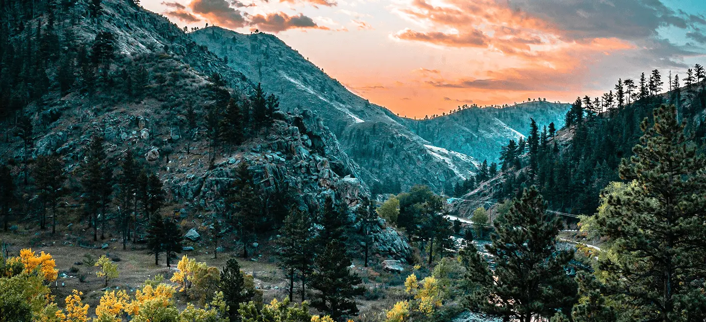Mountain landscape with pine trees and a winding road under a colorful sunset sky.
