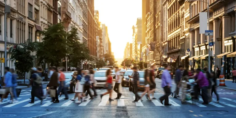 Busy urban crosswalk with blurred pedestrians and cars, surrounded by tall buildings at sunset.