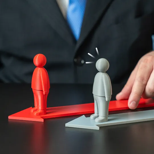 Two plastic figurines, one red and one gray, stand on opposing arrows with a person in a suit in the background.