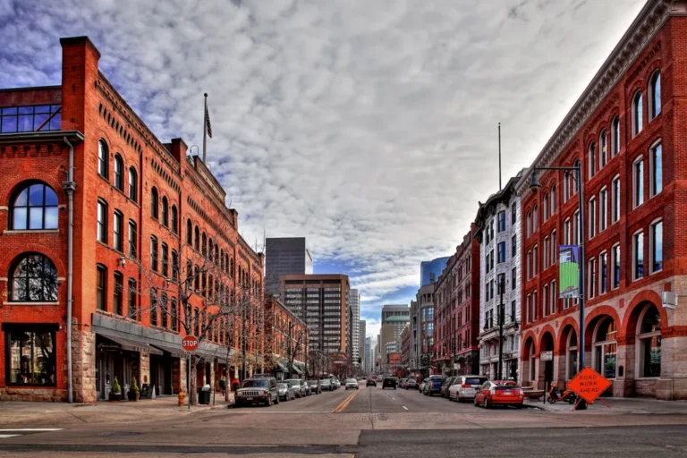 Urban street view with red brick buildings, parked cars, and cloudy sky.
