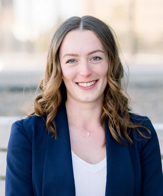 Professional headshot of a smiling woman with wavy brown hair wearing a navy blazer and white top.
