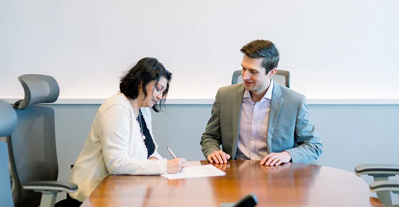Two professionals, a woman in a white blazer and a man in a gray suit, seated at a wooden conference table, with the woman signing a document.