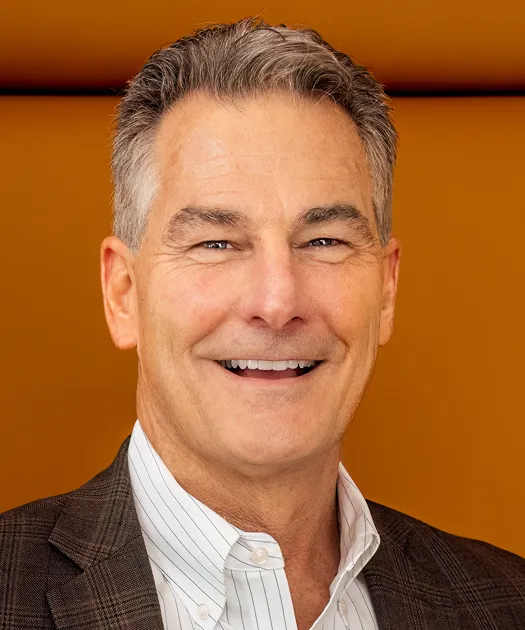 Professional middle-aged man with gray hair, wearing a white striped shirt and brown blazer, smiling against an orange background.