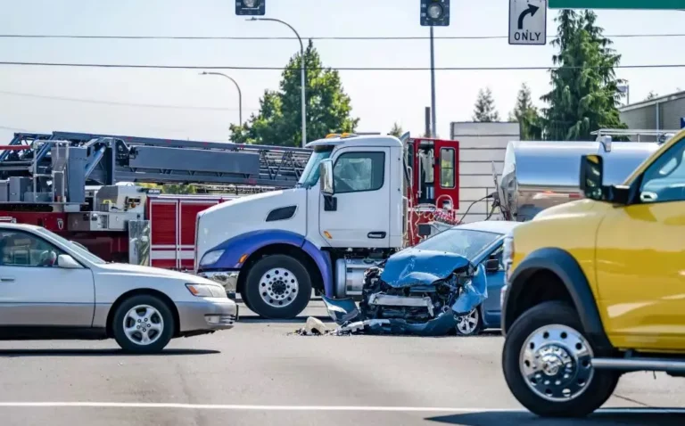 Intersection scene depicting a blue car with severe front-end damage beside a white truck and fire engine.