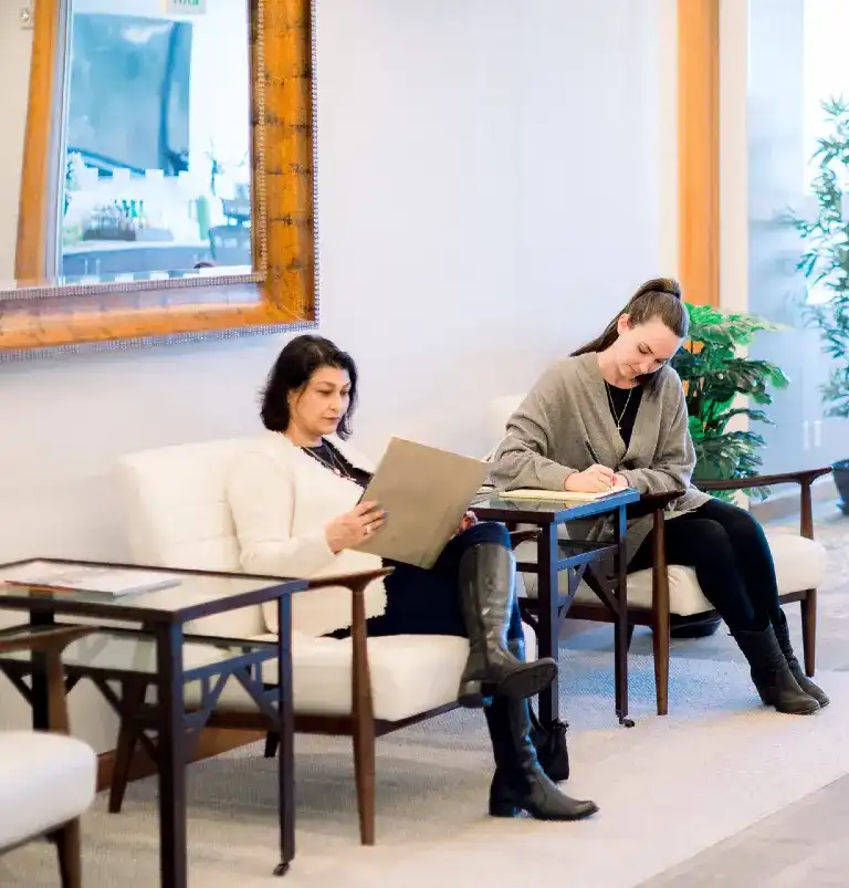 Two women seated in a waiting area, one reading a document and the other writing on a clipboard.