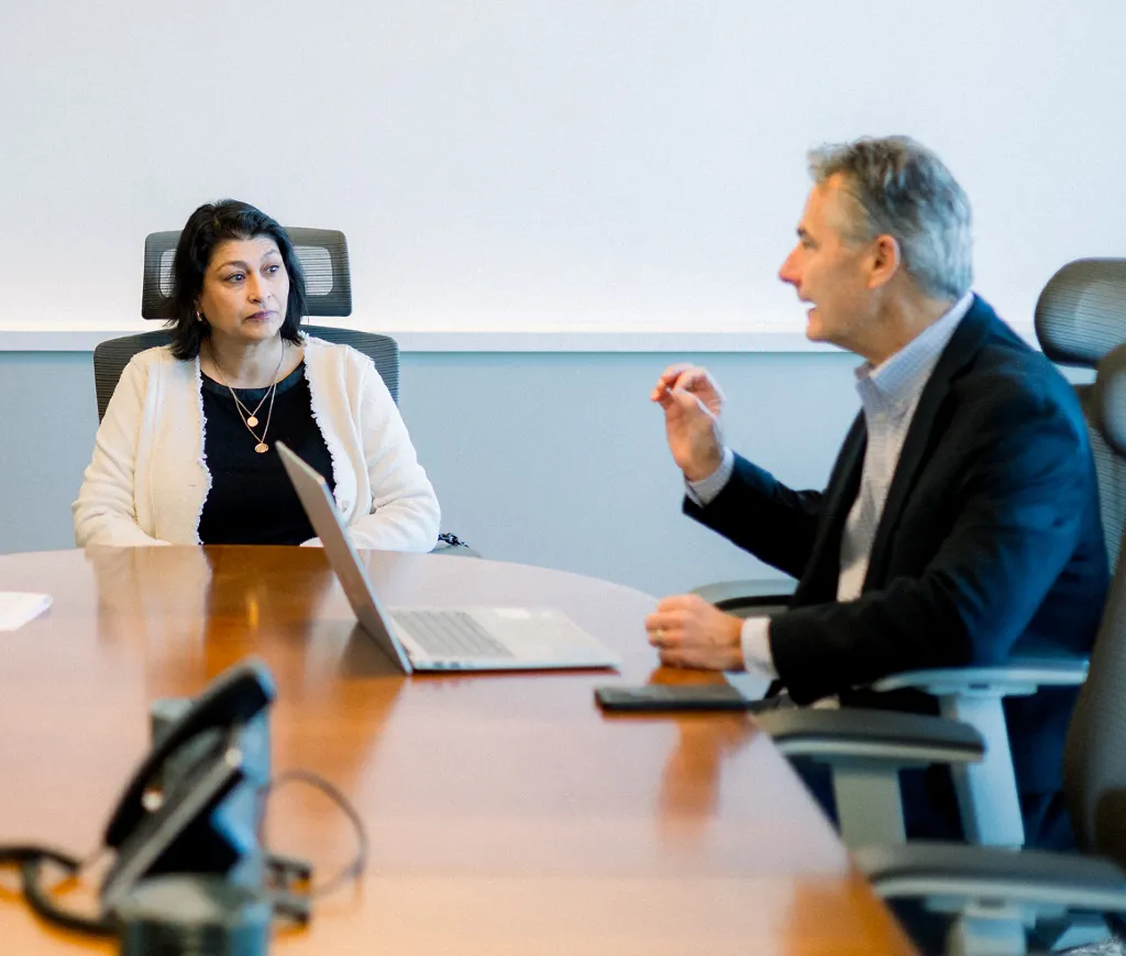 Two professionals engaged in a meeting at a conference table with a laptop and phone.