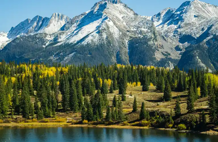 Snow-capped mountain range with dense evergreen forest and reflective lake in foreground.