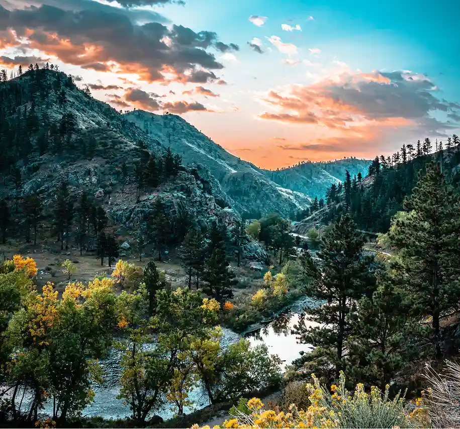 Mountain landscape with a river, pine trees, and a colorful sunset sky.