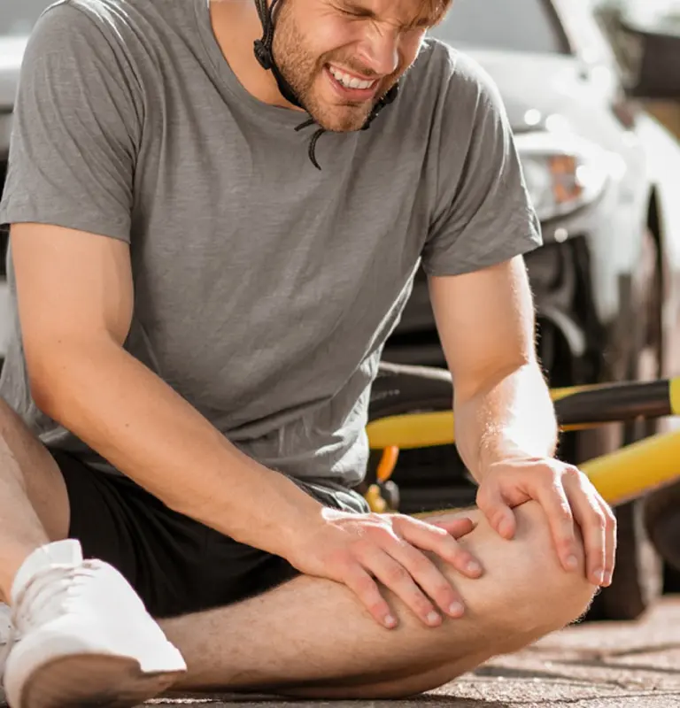 Young man in gray shirt and helmet sitting on ground, holding injured knee in pain.