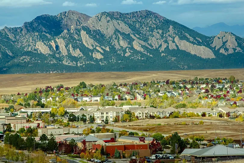 Panoramic view of suburban neighborhood with autumn foliage and large rocky mountains in the background under a partly cloudy sky.