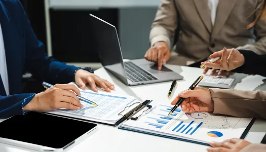 Professionals in business attire analyzing financial charts and documents around a laptop.