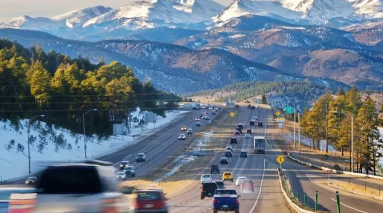 Highway with multiple vehicles driving, surrounded by snow-covered trees and distant snow-capped mountains.