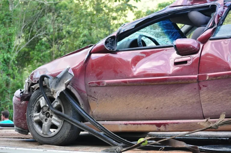 Damaged red car with shattered windshield and bent front left side on flatbed tow truck.