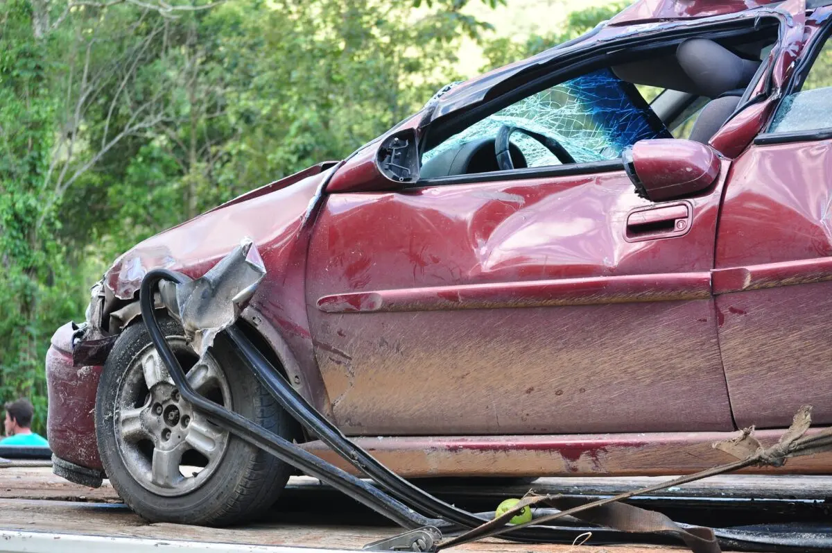 Damaged red car with shattered windshield and bent front left side on flatbed tow truck.