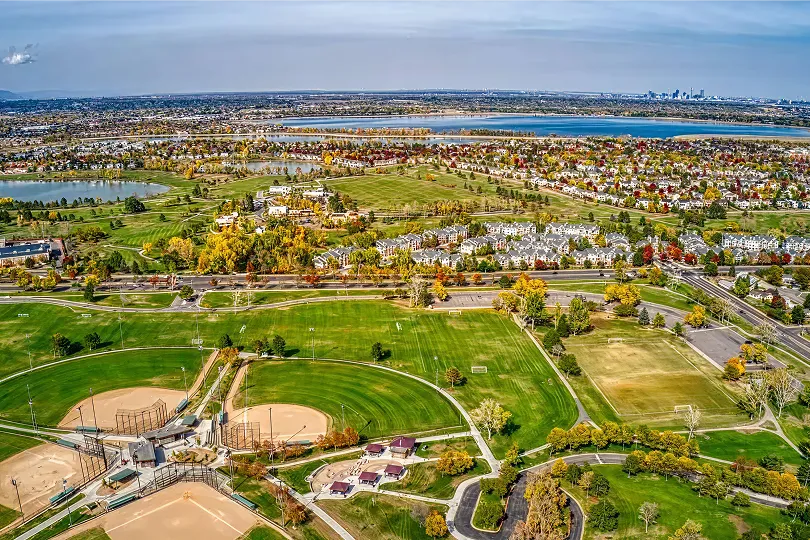 Aerial view of a green park with multiple baseball fields, residential neighborhoods, a large body of water, and a city skyline in the distance.