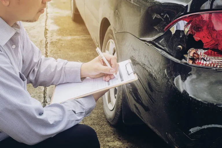 Man in checkered shirt inspecting and writing report on clipboard near damaged black car.