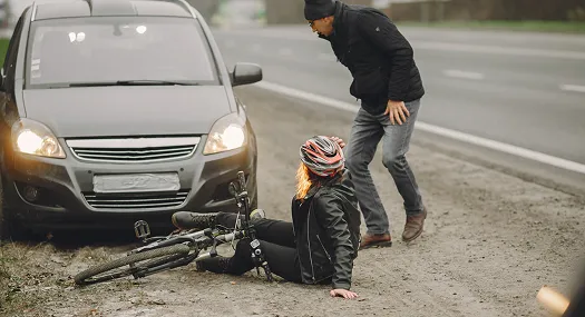 Woman wearing a helmet sitting on the ground next to a fallen bicycle and a concerned man by a black car.