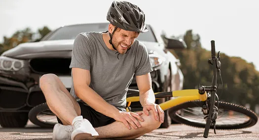 Man wearing a helmet sitting on the ground, holding injured knee beside fallen yellow bicycle and black car.