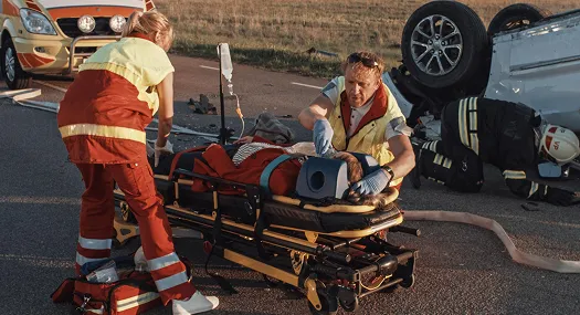 Emergency responders attending to an injured person on a stretcher near an overturned vehicle.