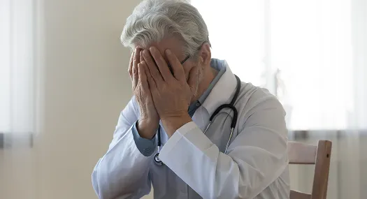 Elderly female doctor in white coat with stethoscope covering face with hands.