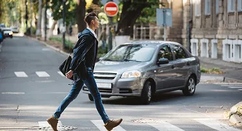 A man wearing a face mask crosses a city street at a crosswalk near a silver car.