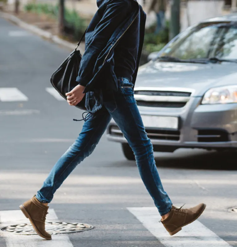 Person wearing blue jeans and brown shoes crossing a street with a silver car in the background.