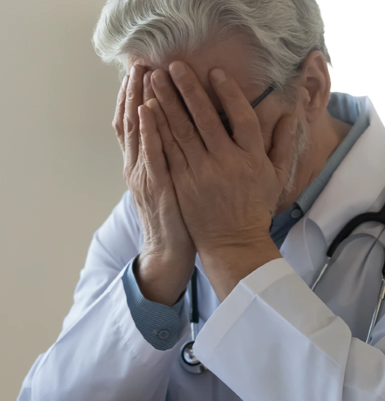 Older male doctor in white coat with stethoscope covering face with hands.