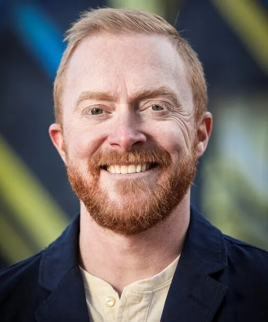 Portrait of a smiling man with red hair and beard, wearing a dark jacket and light shirt.