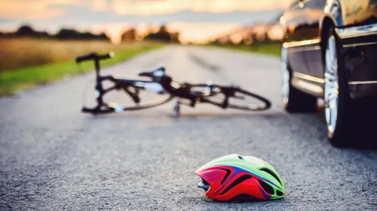 Brightly colored bicycle helmet on road near fallen bicycle and parked car at sunset.