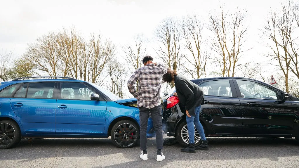 Two individuals inspecting damage after a rear-end collision between a blue and a black car.