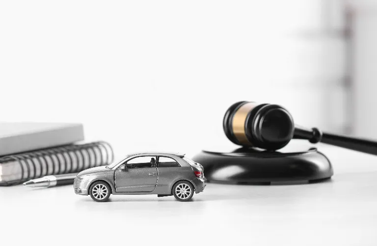 Small gray toy car placed on a white surface next to a black and gold judge's gavel and spiral notebooks.
