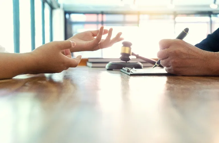 Two individuals at a wooden table, one gesturing with hands, the other writing on a clipboard, with a judge's gavel in the background.