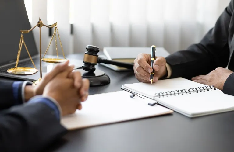 Two professionals in suits at a desk with legal scales, a gavel, and a notebook.