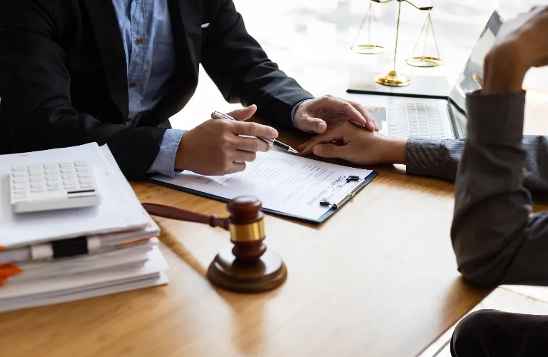 Two professionals at a desk with legal documents, a gavel, and a laptop, one holding the other's hand.