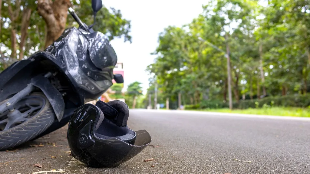 Black motorcycle helmet lying on the road next to a parked black scooter under trees.