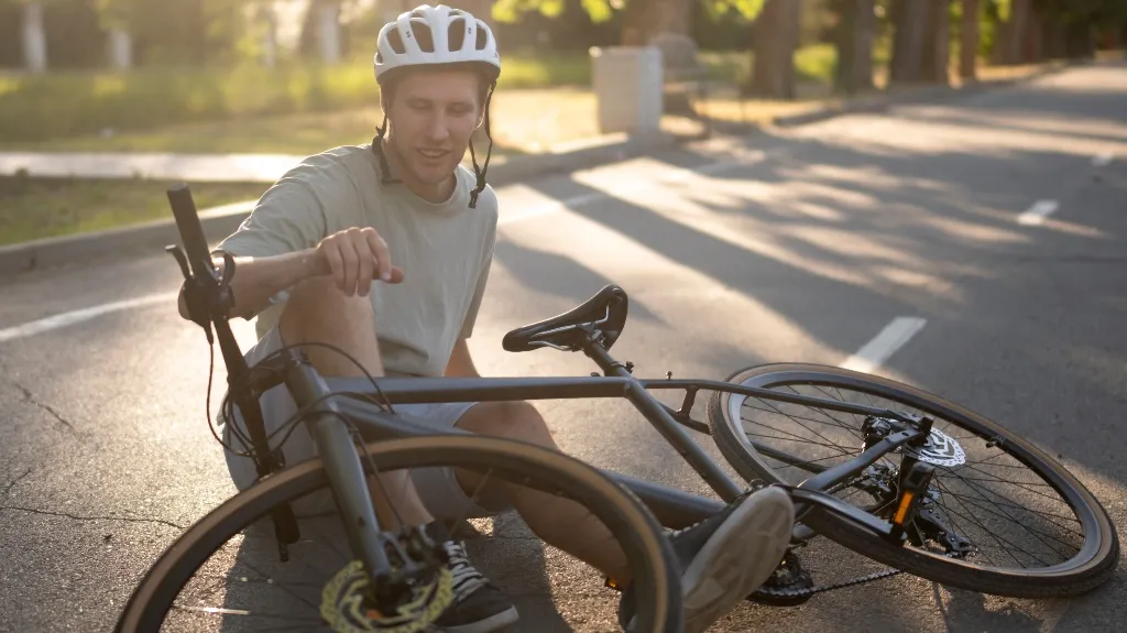 Young man wearing a white helmet sitting on the road next to a fallen black bicycle in daylight.