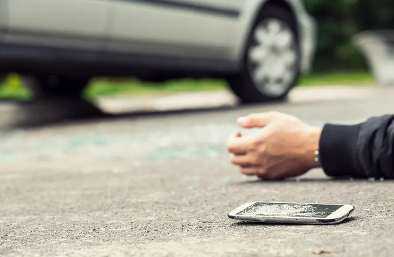 Person lying on pavement near a white car, with a cracked smartphone on the ground.