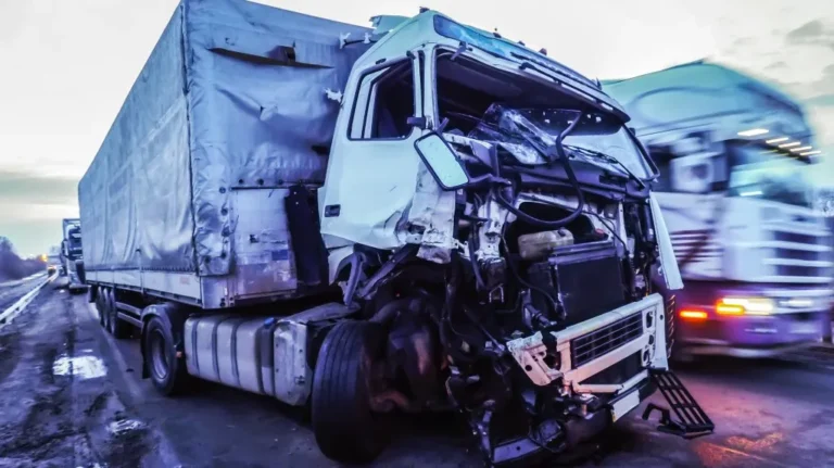 White semi-truck with severe front-end damage parked on roadside during dusk.