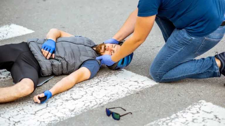 Man wearing a blue helmet and gray vest lies unconscious on road; another person provides first aid.