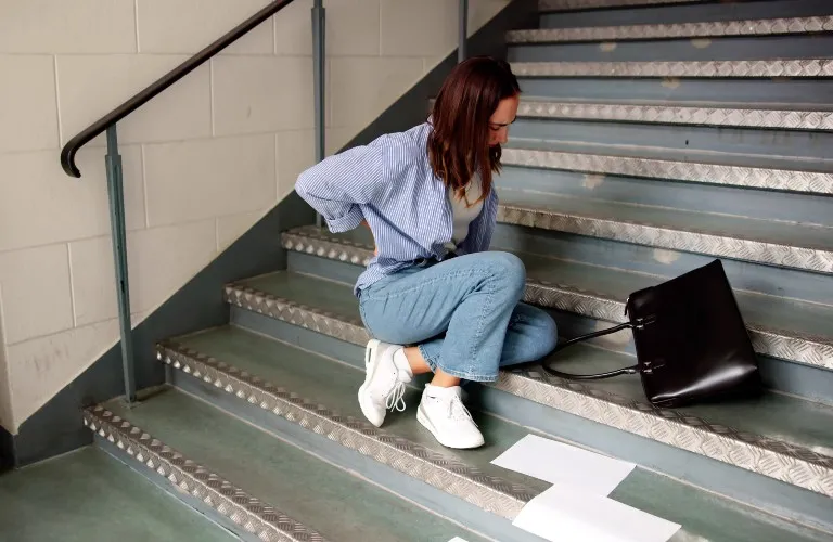 Woman in blue shirt and jeans sitting on gray metal stairs, holding her lower back in pain.