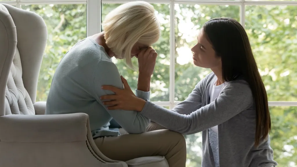 Two women indoors by a large window, one comforting the other who is distressed.