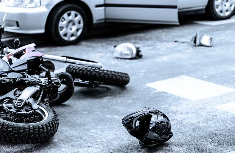 Motorcycle lying on its side on asphalt with two helmets and a silver car nearby.