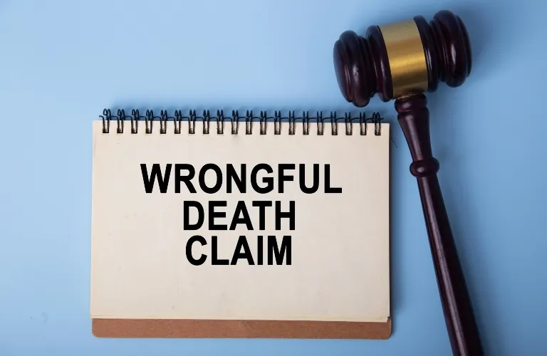 Notepad with "Wrongful Death Claim" text beside a wooden judge's gavel on blue background.