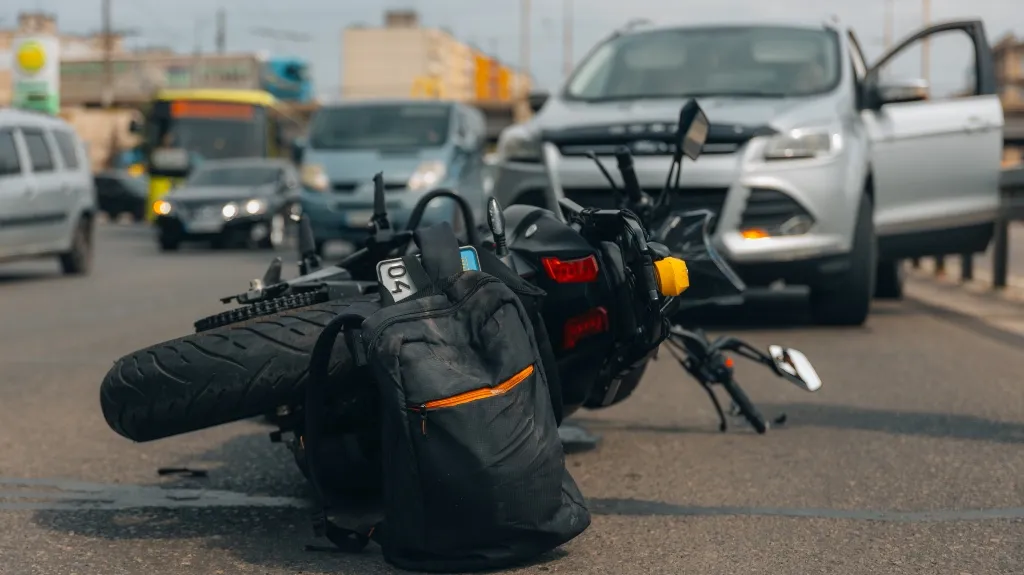 Black motorcycle lying on its side in the street with a black backpack nearby and a white car with open door in the background.