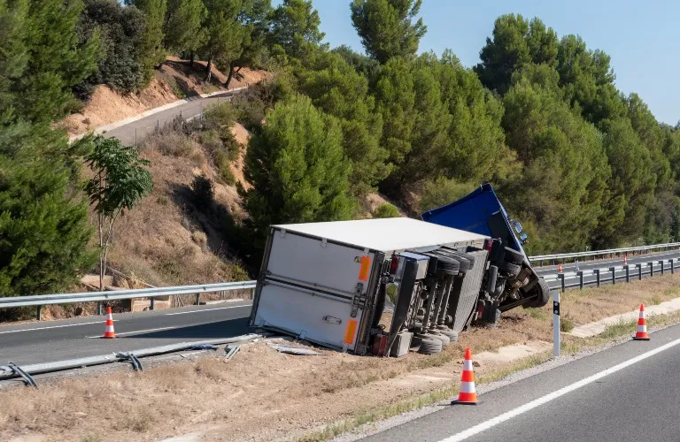 Large blue and white truck overturned on roadside with traffic cones and guardrails.