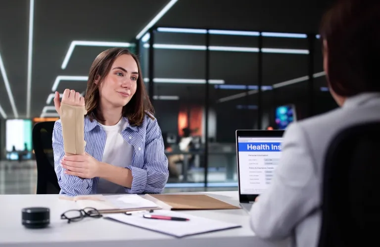 Two women engaged in a professional discussion at an office desk with a laptop displaying health insurance information.