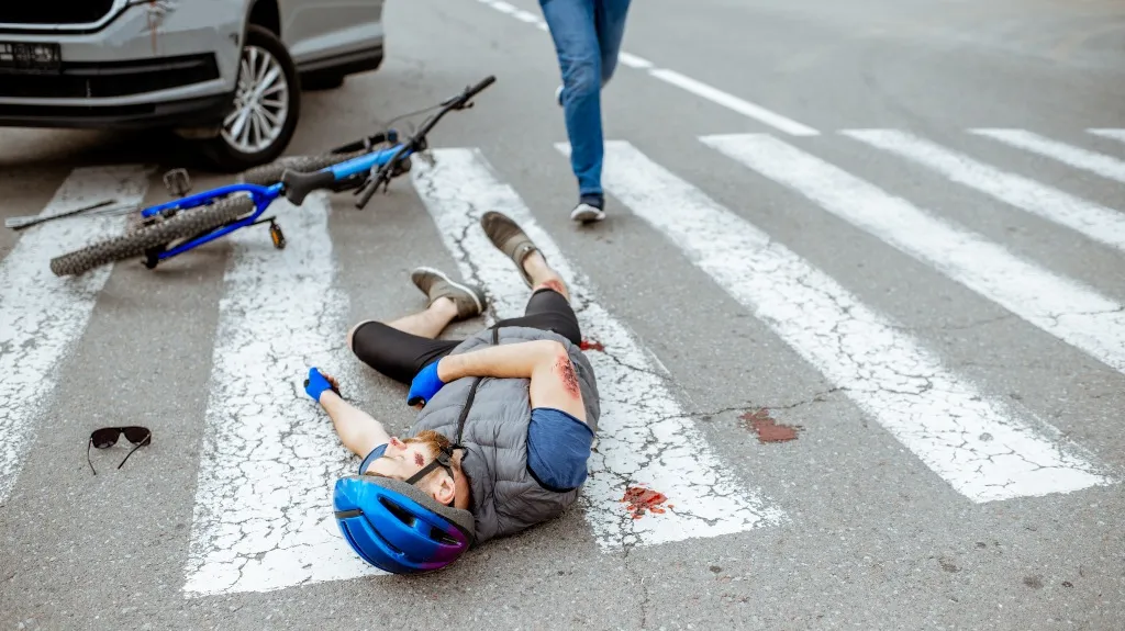 Man wearing a blue helmet lying injured on a crosswalk beside a blue bicycle and a white car.