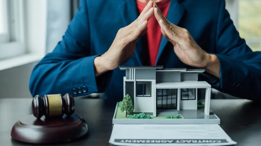 Person in blue blazer with hands forming a roof gesture over a house model and contract agreement.