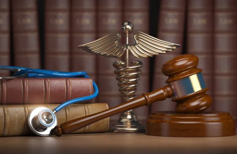 Wooden judge's gavel, medical caduceus symbol, stethoscope, and law books on a desk.