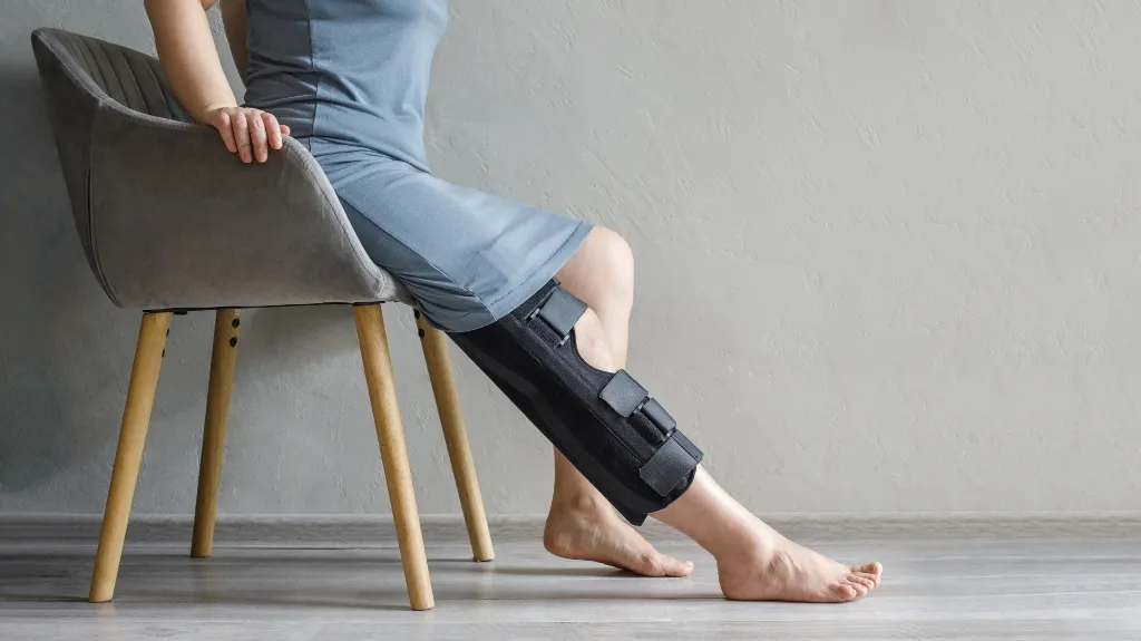 Person seated on gray chair wearing a gray dress and a black leg brace on right leg.